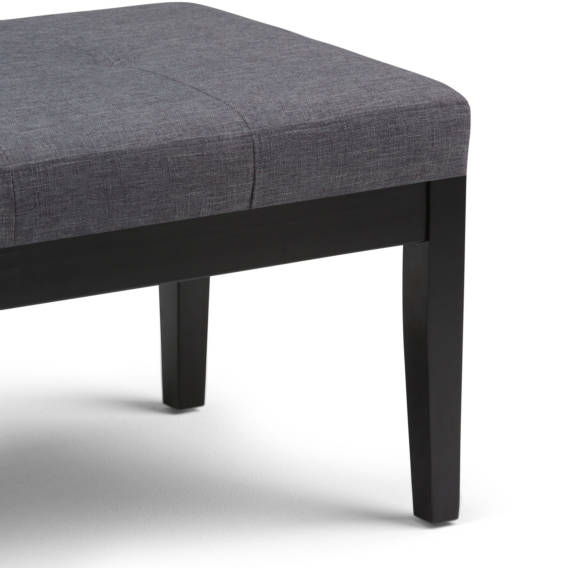 The Lacey Upholstered Tufted Ottoman Bench features a cushioned gray fabric seat and dark wooden legs, shown in close-up against a white background.