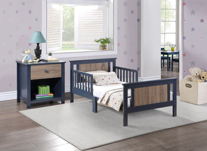 A child’s bedroom with a blue and wood-tone Connelly Reversible Panel Toddler Bed, gray rug, nightstand with lamp and books, star wallpaper, shuttered window, and a box of stuffed animals on the wooden floor.