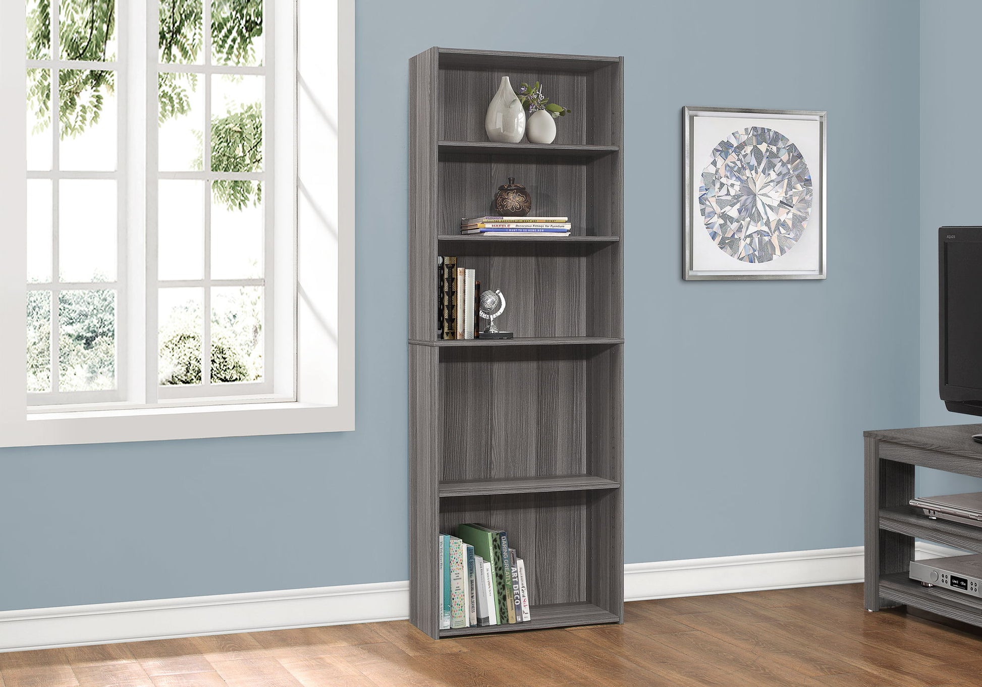 A tall gray 6-tier transitional bookshelf stands against a light blue wall near a window, beside a framed picture and TV stand, holding books and decor on the wooden floor—perfect for office or home use.