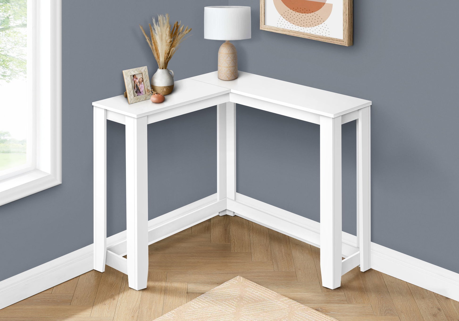 A Corner Accent Console Table for Entryway with a lamp, framed photo, vase of dried plants, and decorative object stands against a gray wall with wood flooring and a window nearby. Its unique contemporary design enhances the space.