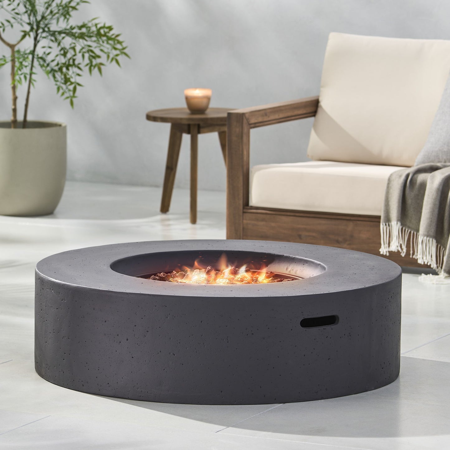 The Modern Lightweight Concrete Outdoor Circular Fire Pit 50,000 Btu, featuring a smooth gray finish and bright flame, is on a patio beside a beige cushioned sofa, wooden table with candle, and potted plant in the background.