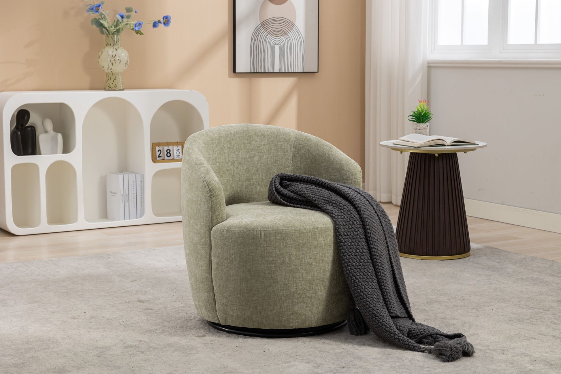 A Chenille Fabric Swivel Accent Armchair Barrel Chair with a dark gray knit blanket draped over the arm sits on a beige rug. Behind it are a white shelf, a round side table with an open book, and framed art on a light tan wall.