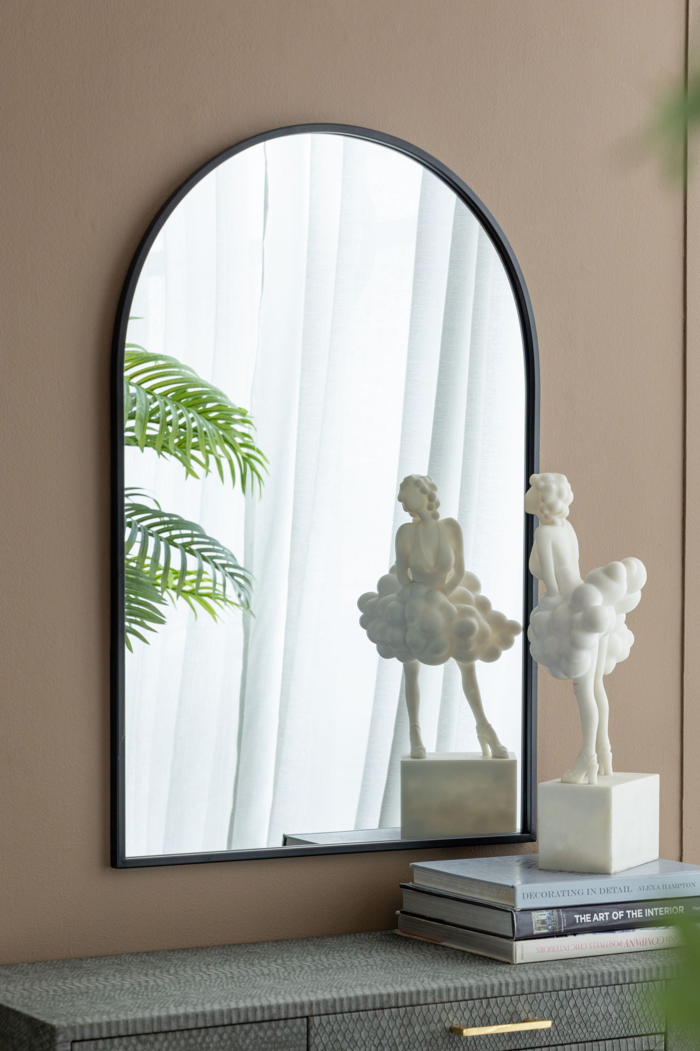 An Arched Mirror With Metal Frame reflects a leafy plant and sheer curtains. In front, a cloud-shaped statue sits on stacked books on a textured surface, making it perfect for living rooms, bedrooms, or entryways.