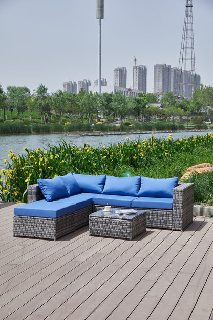 The Patio Sofa With Cushions, featuring bright blue cushions, is set on a wooden deck by a pond with tall green plants and yellow flowers. City buildings and a metal tower can be seen in the distance.