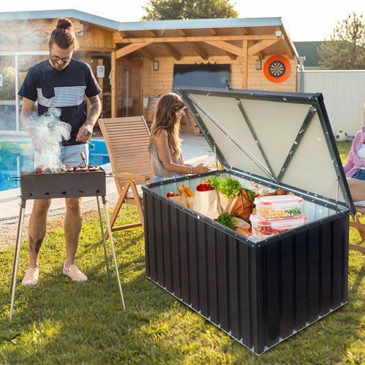 A man grills outdoors as a woman sits nearby, with a wooden shed and pool in the background. An open Metal Storage Box Store Small - Black, filled with groceries and vegetables, is placed on the grass.