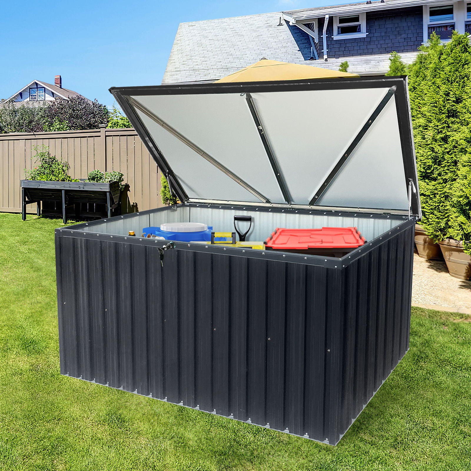 The Metal Storage Box Store Large Out Door Use - Black sits on a green lawn near a house and garden, its lid open to reveal storage for items such as a shovel, red container, and blue bucket.