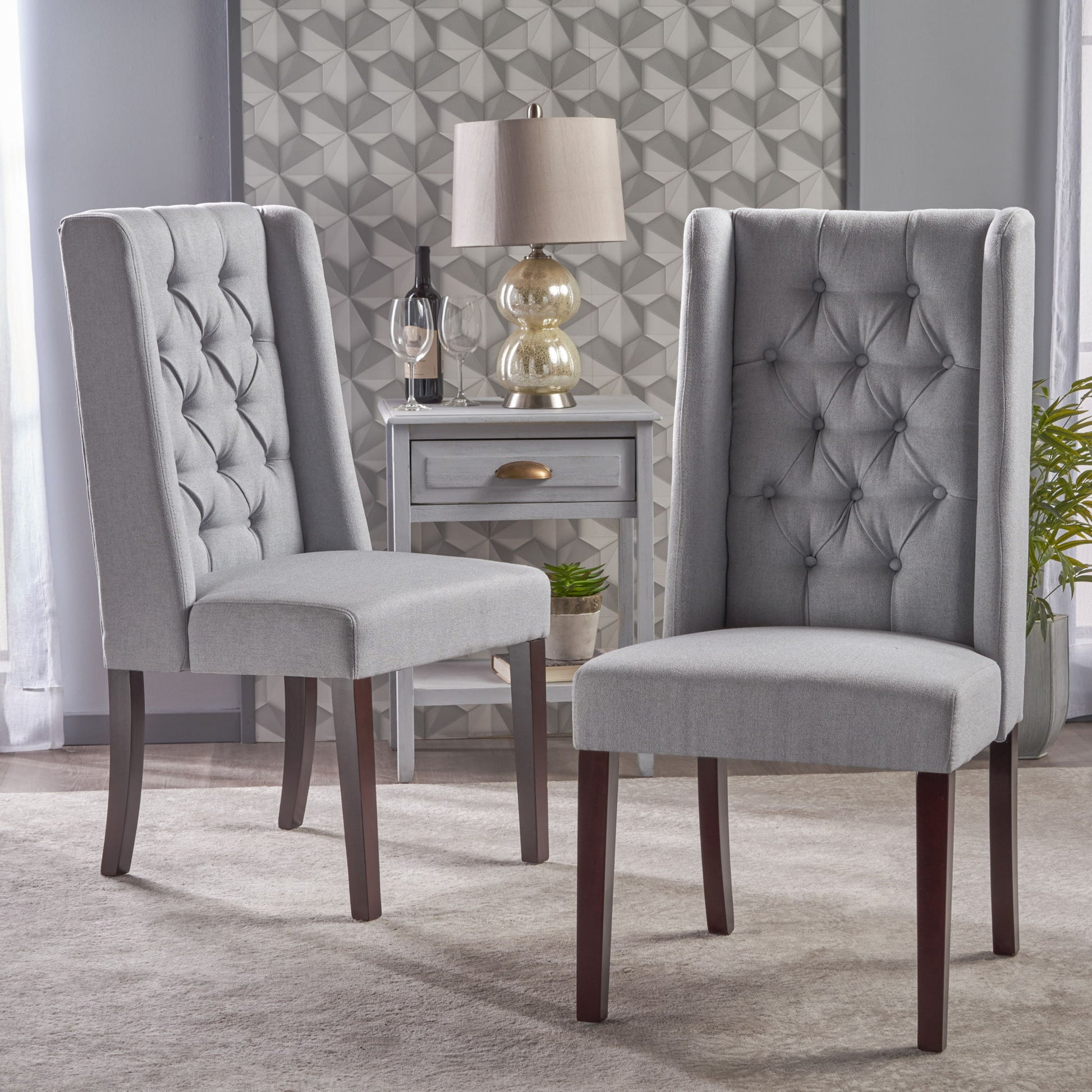 Two Sophisticated Tufted Dining Chairs With High Back Design are placed on a rug in front of a small gray table with a drawer, lamp, plant, and decor against a patterned accent wall.