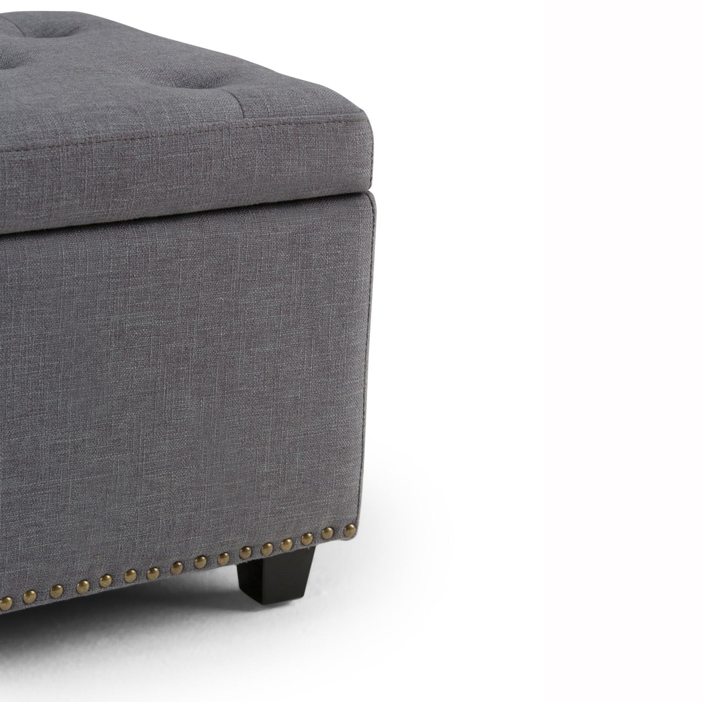 Close-up of the Hamilton Upholstered Storage Ottoman's corner, featuring a gray tufted top, black wood legs, and brass nailhead trim along the bottom edge.