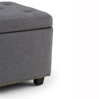 Close-up of the Hamilton Upholstered Storage Ottoman's corner, featuring a gray tufted top, black wood legs, and brass nailhead trim along the bottom edge.