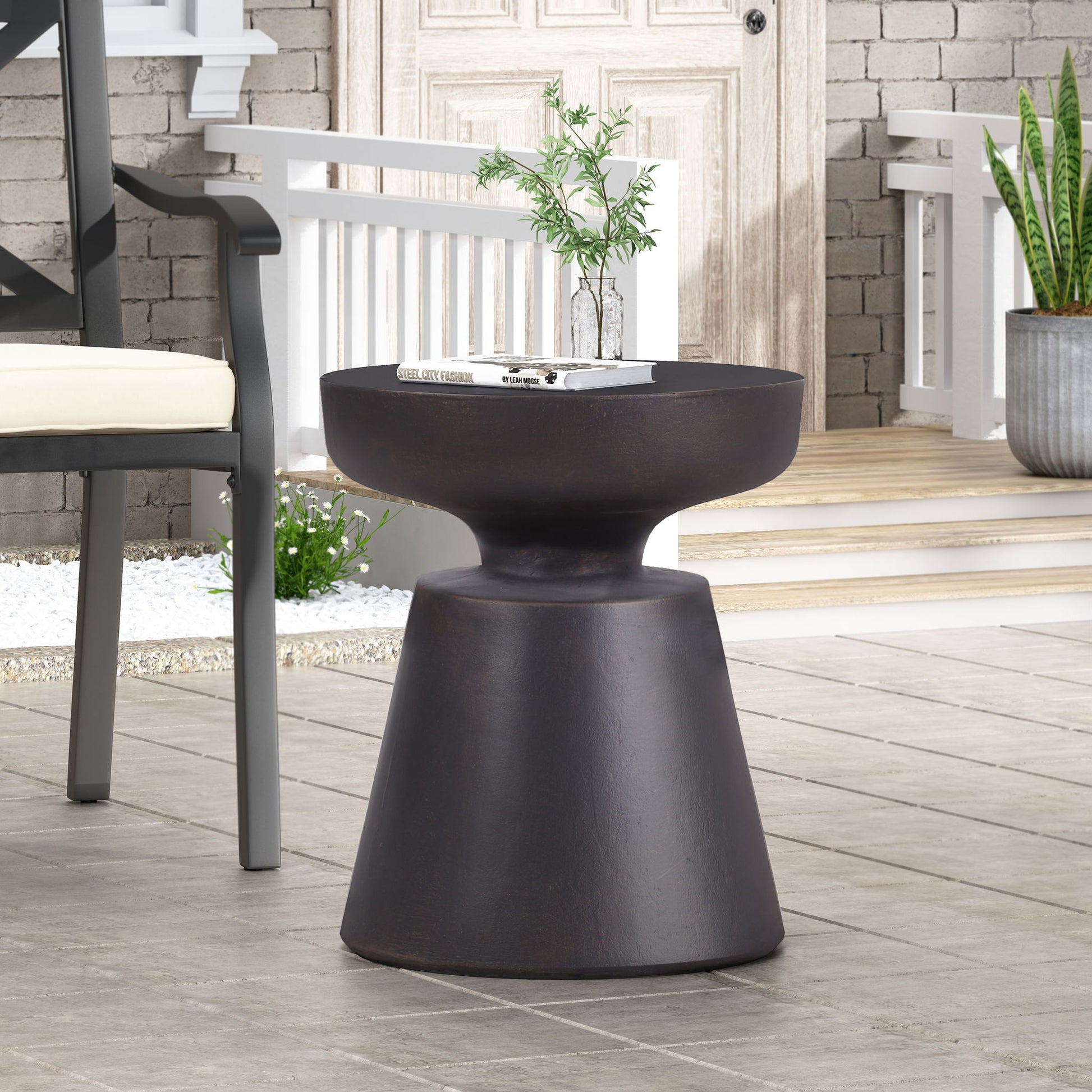The Outdoor Lightweight Concrete End Table With Half Sphere Top - Copper, featuring a modern dark brown finish and rounded top with tapered base, sits on a tiled patio. A glass vase with green branches and a book rest on it; a chair and plants are behind.
