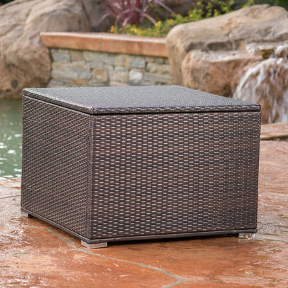 The Santa Rosa - Storage Table, a dark brown outdoor wicker box, is placed on a stone patio near a pond with rocks and a small waterfall in the background.