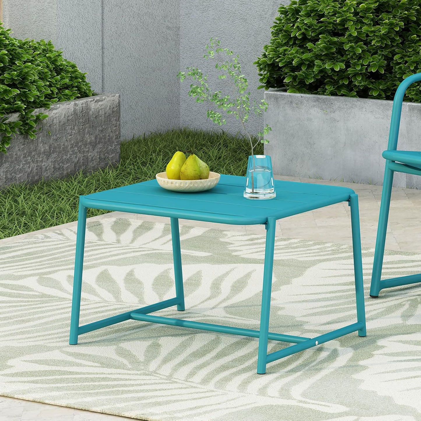 The Conrad - Side Table Outdoor in teal sits on a leaf-patterned rug, topped with a bowl of pears and a sprig-filled glass, amid green shrubs and concrete planters.
