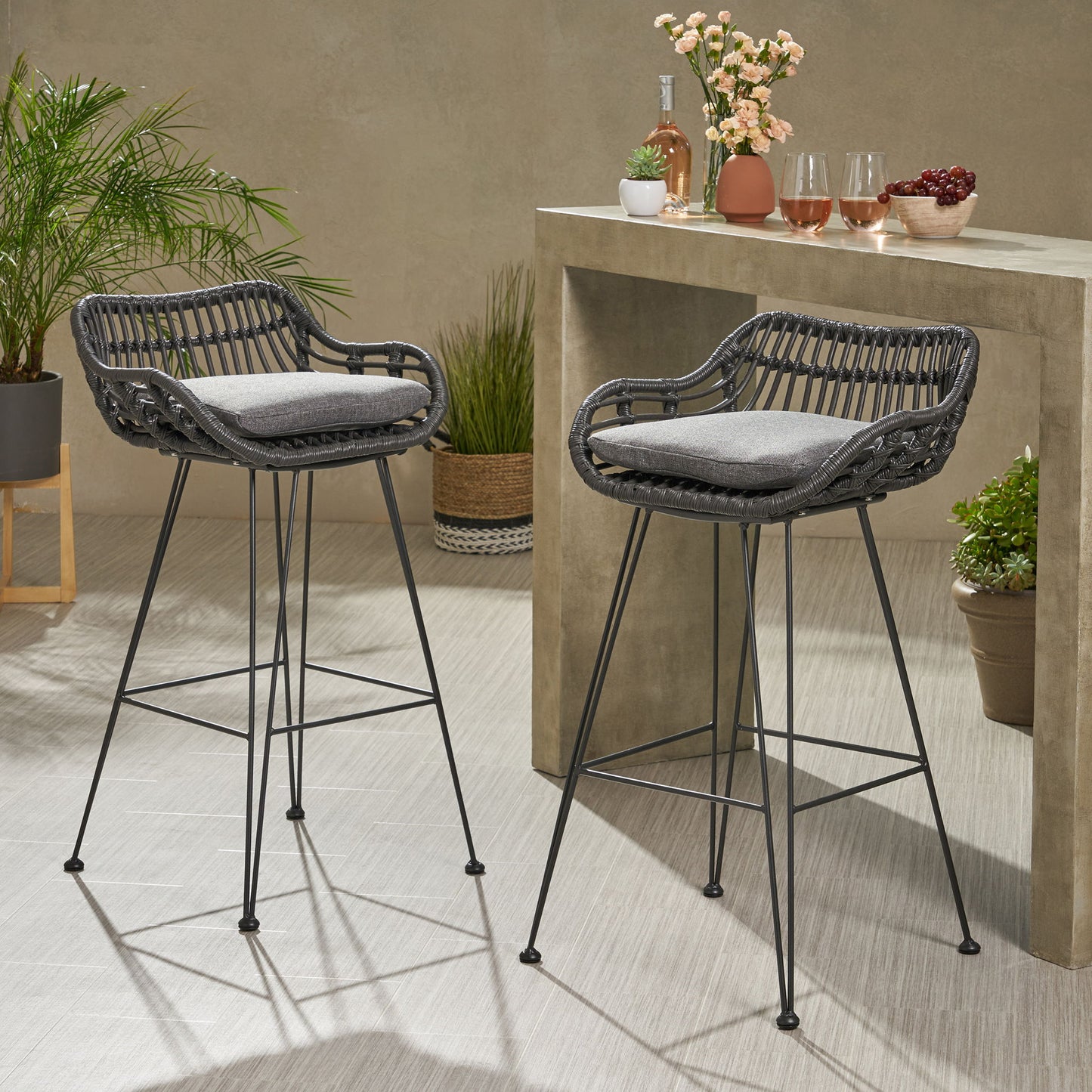 Two Outdoor Rattan Bar Chairs with metal legs and gray cushions sit by a stone bar table topped with potted plants, drinks, glasses, and cherries, creating a stylish modern scene for indoor or patio use.