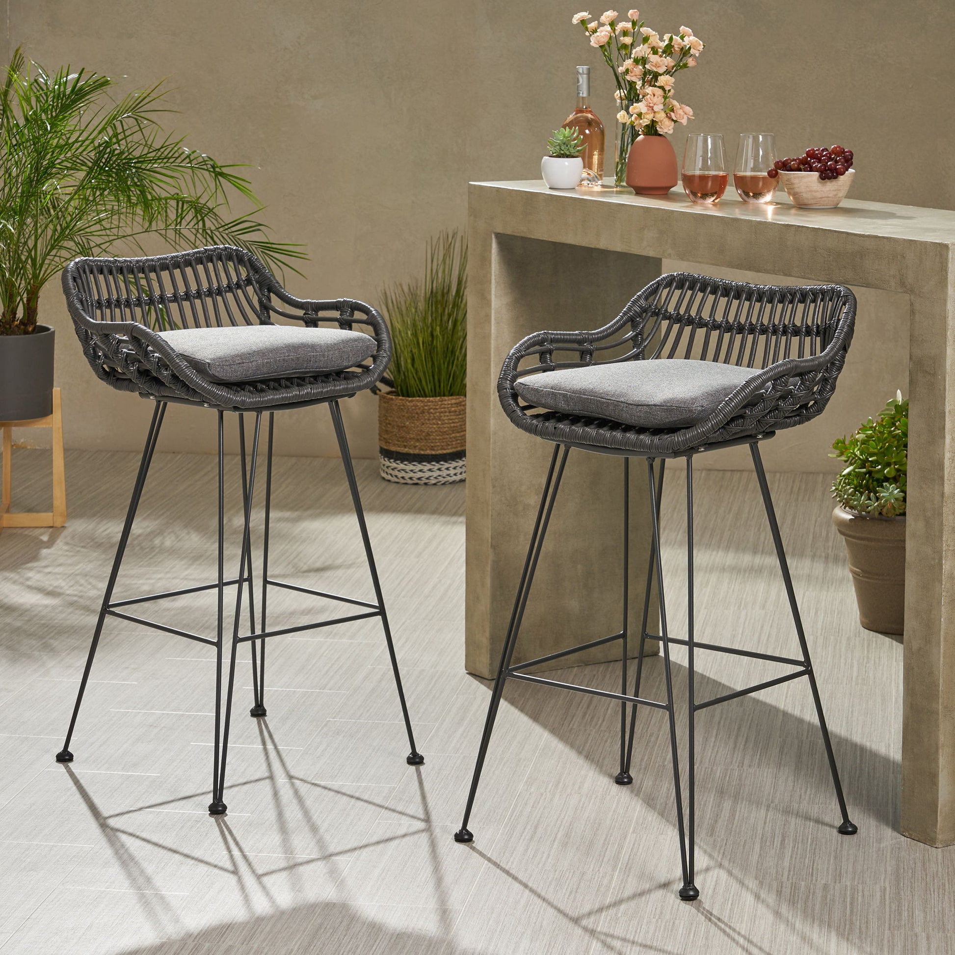 Two Outdoor Rattan Bar Chairs with metal legs and gray cushions sit by a stone bar table topped with potted plants, drinks, glasses, and cherries, creating a stylish modern scene for indoor or patio use.