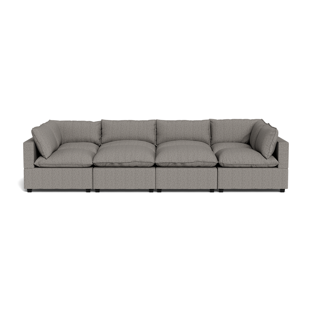 The Kova Pillow Cushion Grand Pit is a modern gray sectional sofa with thick seat and back cushions, four seating sections, and armrests on both ends, shown from the front against a black background.