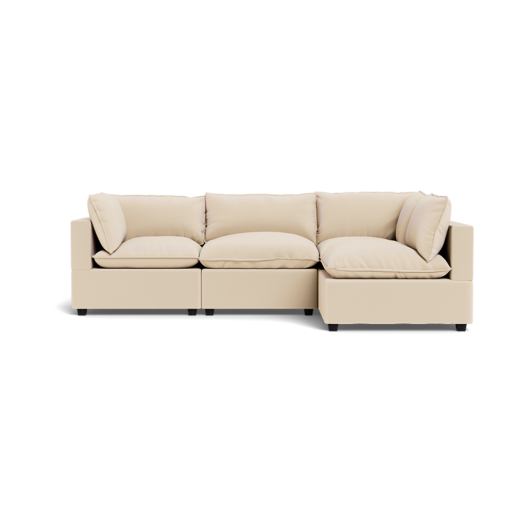 The Kova Pillow Cushion L-Shape is a modern, light beige sectional sofa with three seat cushions and four back pillows, shown from the front on a plain white background.