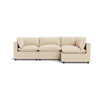 The Kova Pillow Cushion L-Shape is a modern, light beige sectional sofa with three seat cushions and four back pillows, shown from the front on a plain white background.
