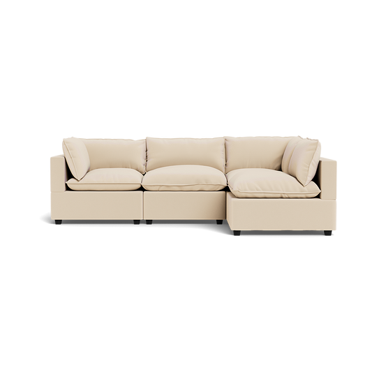 The Kova Pillow Cushion L-Shape is a modern, light beige sectional sofa with three seat cushions and four back pillows, shown from the front on a plain white background.
