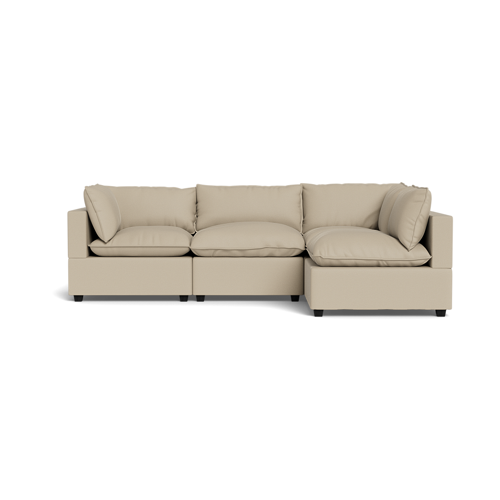 The Kova Pillow Cushion L-Shape is a modern beige sectional sofa with three seat cushions, four back cushions, wide armrests, and a low profile, displayed against a plain black background.