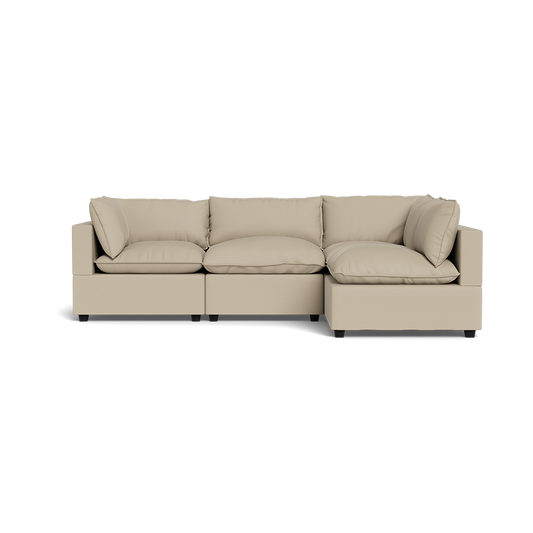 The Kova Pillow Cushion L-Shape is a modern beige sectional sofa with three seat cushions, four back cushions, wide armrests, and a low profile, displayed against a plain black background.