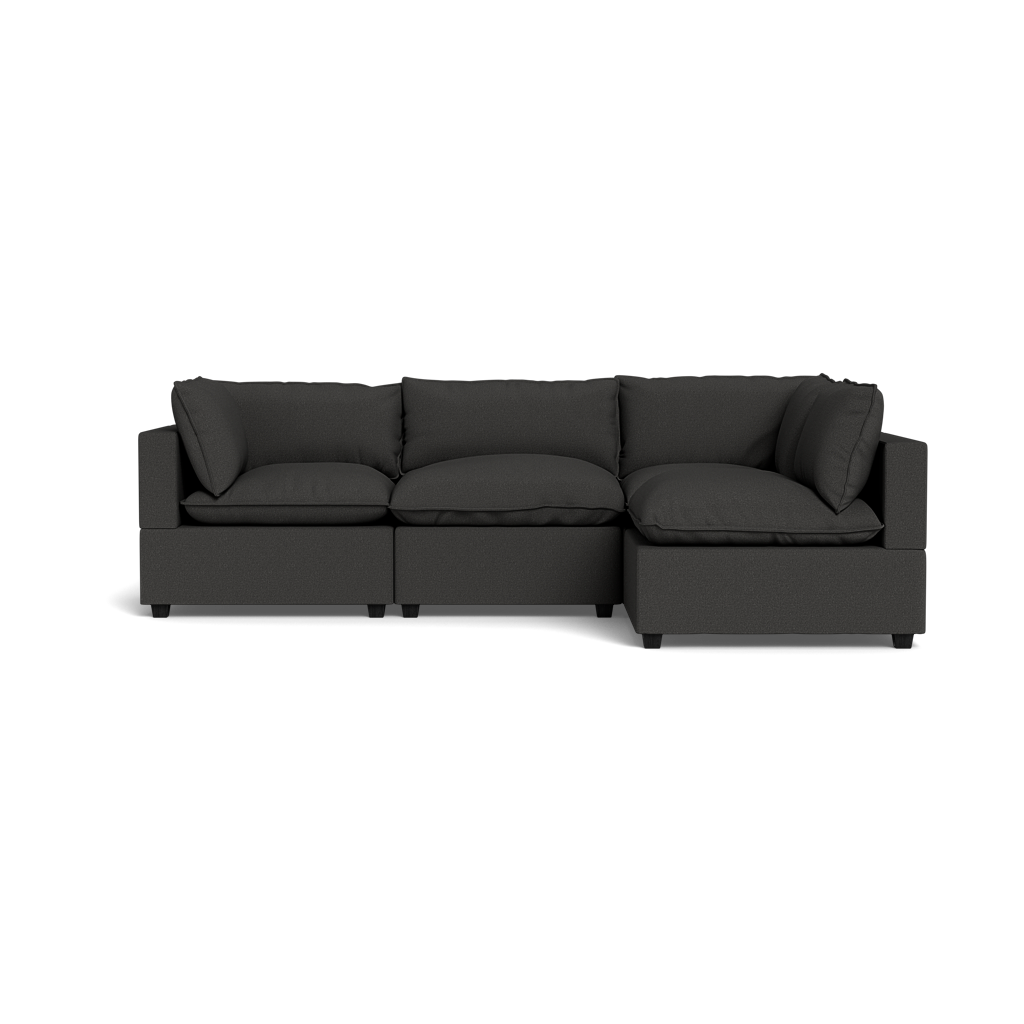 The Kova Pillow Cushion L-Shape is a modern dark gray sectional with three seat cushions, three back cushions, and wide armrests, displayed against a plain black background.