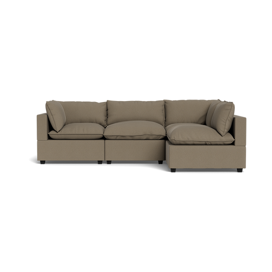 The Kova Pillow Cushion L-Shape is a modern, three-seater sectional sofa in light brown fabric with wide armrests and plush cushions, shown from the front against a plain black background.
