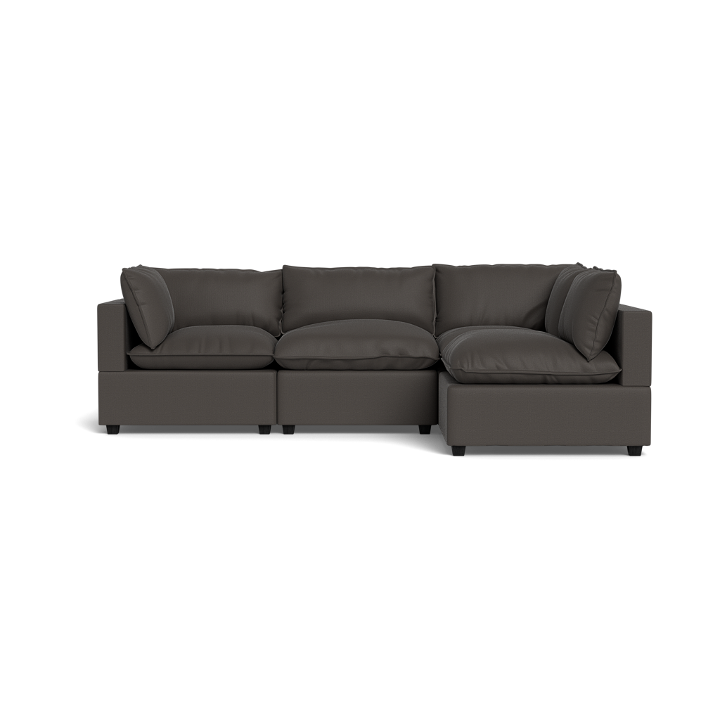 The Kova Pillow Cushion L-Shape is a modern dark gray sectional sofa with plush cushions and wide armrests, shown from the front against a black background.