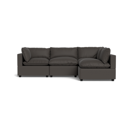 The Kova Pillow Cushion L-Shape is a modern dark gray sectional sofa with plush cushions and wide armrests, shown from the front against a black background.