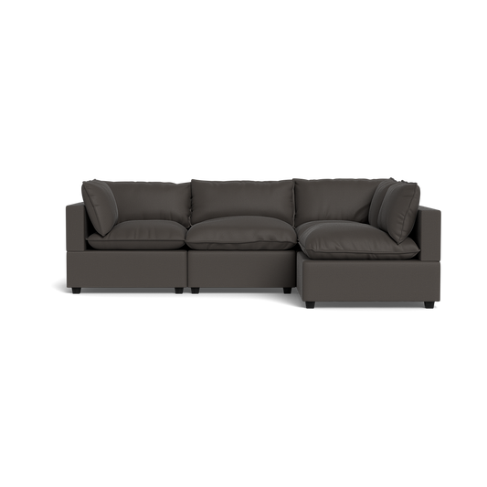 The Kova Pillow Cushion L-Shape is a modern dark gray sectional sofa with plush cushions and wide armrests, shown from the front against a black background.