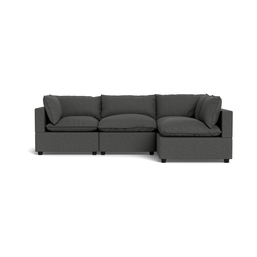 The Kova Pillow Cushion L-Shape is a dark gray, three-seat sectional sofa with wide armrests and plush cushions, shown from the front against a black background.