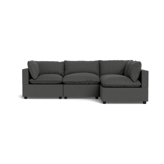 The Kova Pillow Cushion L-Shape is a dark gray, three-seat sectional sofa with wide armrests and plush cushions, shown from the front against a black background.