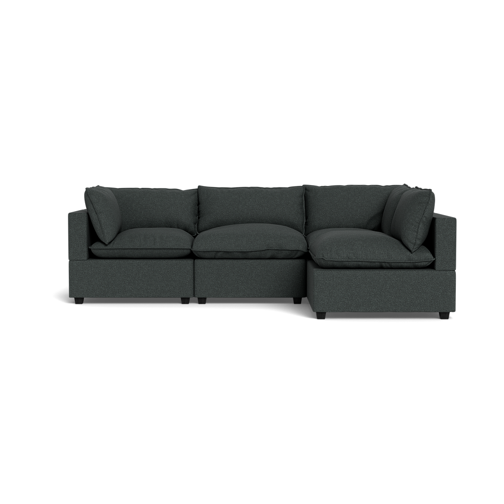 The Kova Pillow Cushion L-Shape is a dark gray sectional sofa with three seat cushions, backrest pillows, and armrests at both ends. It features a modern, boxy design and is shown from the front against a plain black background.