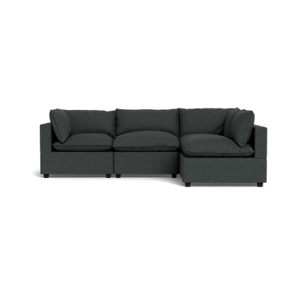 The Kova Pillow Cushion L-Shape is a dark gray sectional sofa with three seat cushions, backrest pillows, and armrests at both ends. It features a modern, boxy design and is shown from the front against a plain black background.