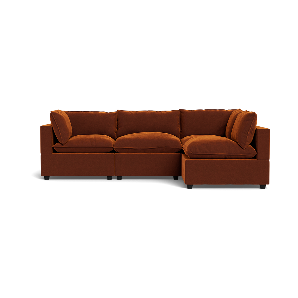 The Kova Pillow Cushion L-Shape is a modern burnt orange sectional sofa with three seat cushions, plush backrest pillows, and a right-side chaise lounge, shown against a black background.