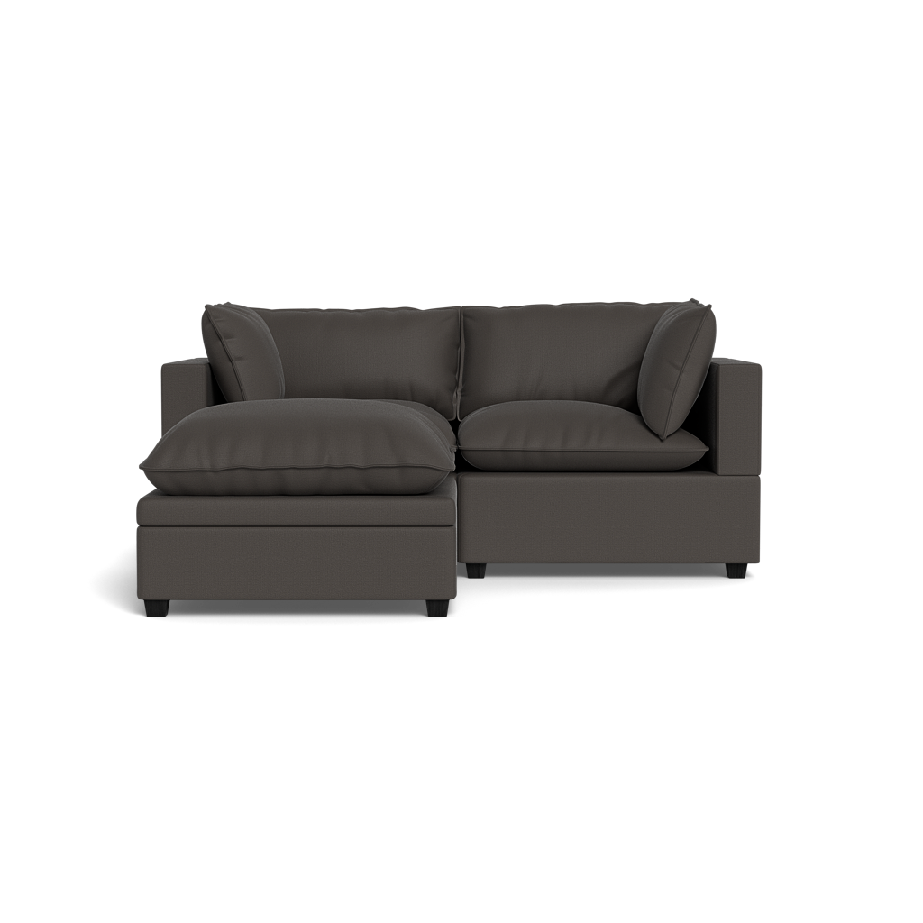 The Kova Pillow Cushion Sofa 86" + Ottoman features plush seat and back cushions, square armrests, and a left-side chaise lounge in dark gray, displayed against a plain black background.