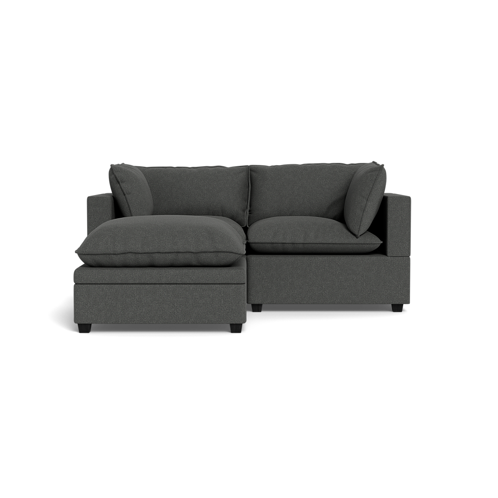 The Kova Pillow Cushion Sofa 86" + Ottoman is a dark grey sectional with wide cushions and armrests, featuring a left-side chaise lounge, displayed against a plain black background.