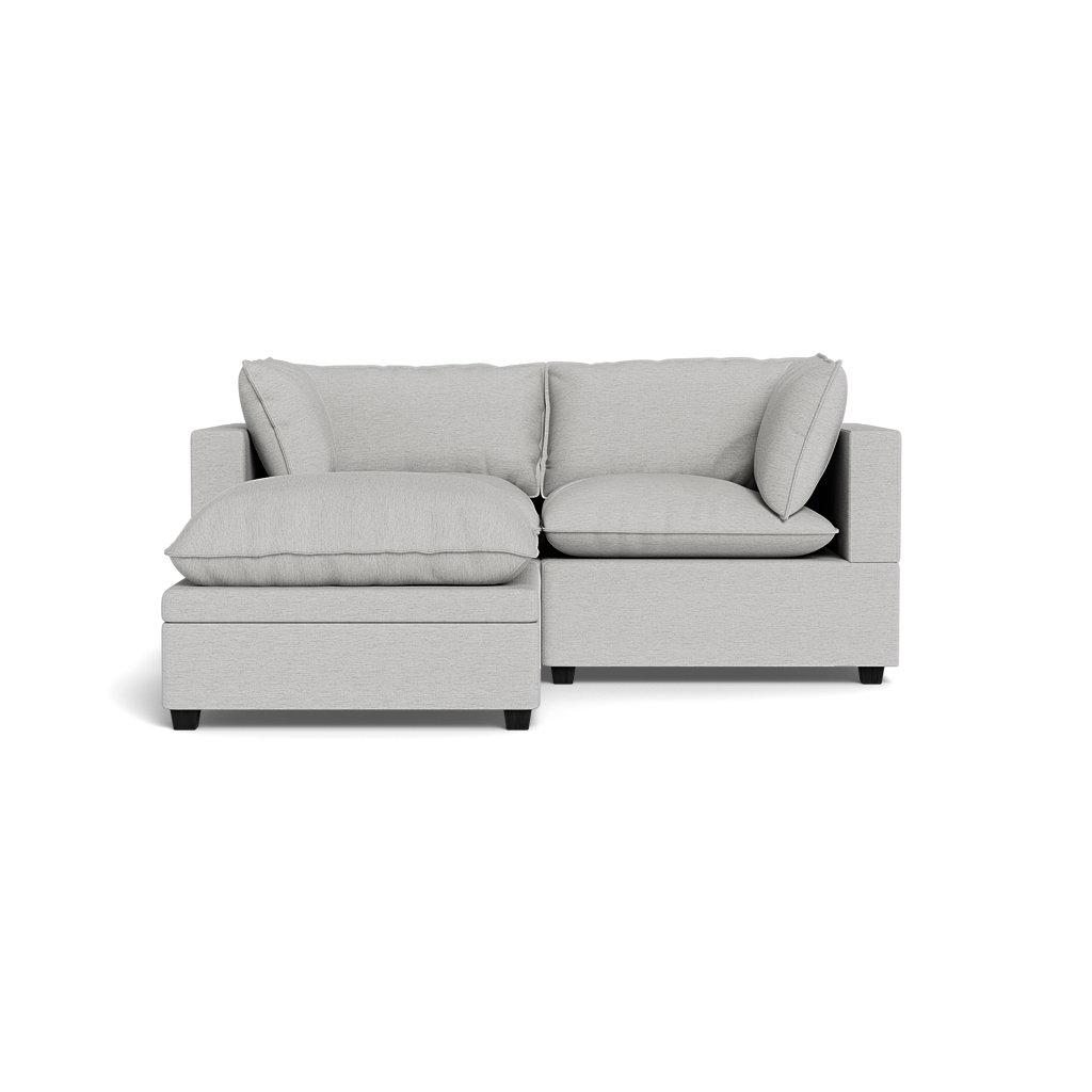 The Kova Pillow Cushion Sofa 86" with Ottoman features plush cushions and an L-shaped design in modern light gray, shown from the front against a solid black background.