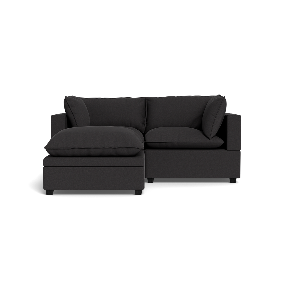The Kova Pillow Cushion Sofa 86" with ottoman features a modern dark gray design, plush cushions, and a left-side extended chaise lounge, beautifully displayed against a plain black background.