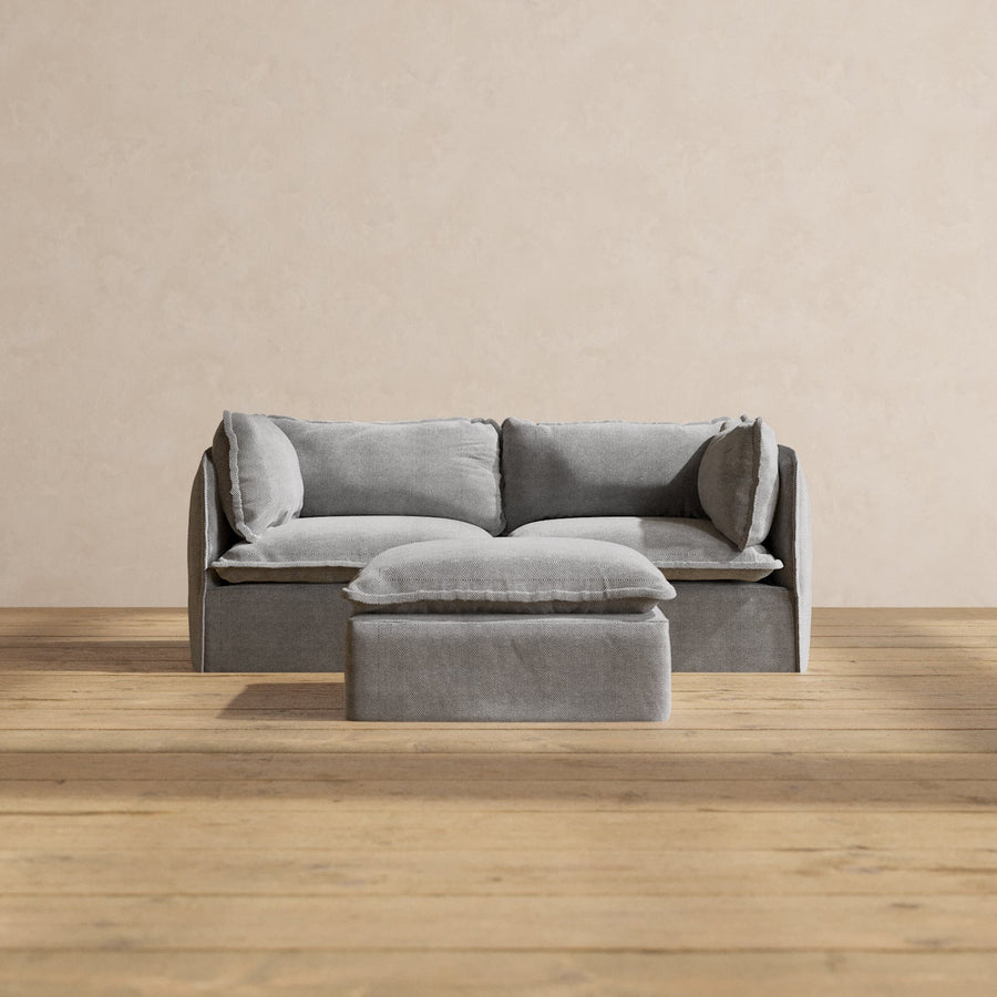 A chic gray Modular Performance 2-Seater with coordinated cushions is set against a beige backdrop. In front of the sofa, a square gray ottoman complements the scene, resting on a light wooden floor. The arrangement exudes a minimalist and tranquil ambiance.