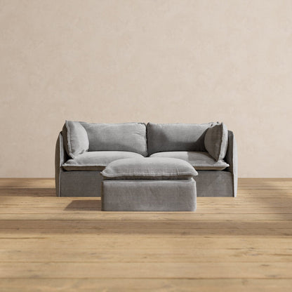 The Modular Performance 2-Seater in minimalist gray, complete with matching pillows, is centered against a beige wall. The set includes a small gray ottoman positioned in front of the sofa. This arrangement sits atop light wooden flooring, creating a simple and modern aesthetic.