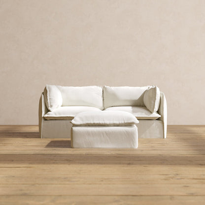A Modular Performance 2-Seater in a minimalist beige design with matching cushions is set on a wooden floor against a plain beige wall. In front of the sofa sits the matching rectangular ottoman with a soft cushion that enhances the neutral color scheme.