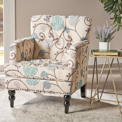 The Vitality Club Chair, featuring beige upholstery with blue and gray floral patterns, sits on a light rug beside a modern table topped with lavender. Sunlight streams in from a window, highlighting a green plant in the background.