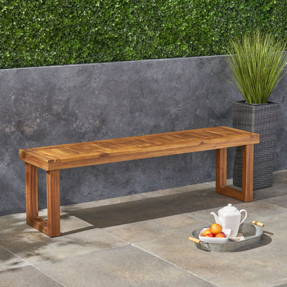 The Nestor - Bench, featuring a simple modern wooden design, sits on a stone patio against a gray wall with a green hedge. A metal tray with teapot, cup, and fruit is beside the bench near a tall potted plant.
