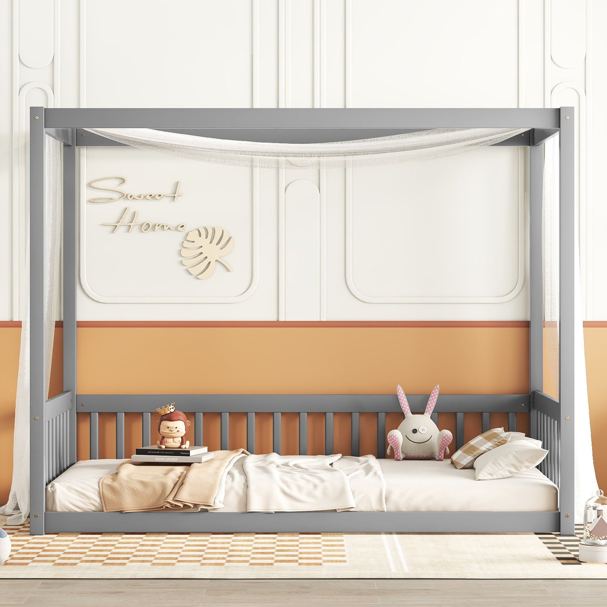 A gray Canopy Frame Floor Bed With Fence and guardrails features a white net, soft bedding, plush toys, pillows, and sits by a peach and white wall with “Sweet Home” decor. A checkered rug lies on the floor.
