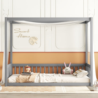 A gray Canopy Frame Floor Bed With Fence and guardrails features a white net, soft bedding, plush toys, pillows, and sits by a peach and white wall with “Sweet Home” decor. A checkered rug lies on the floor.