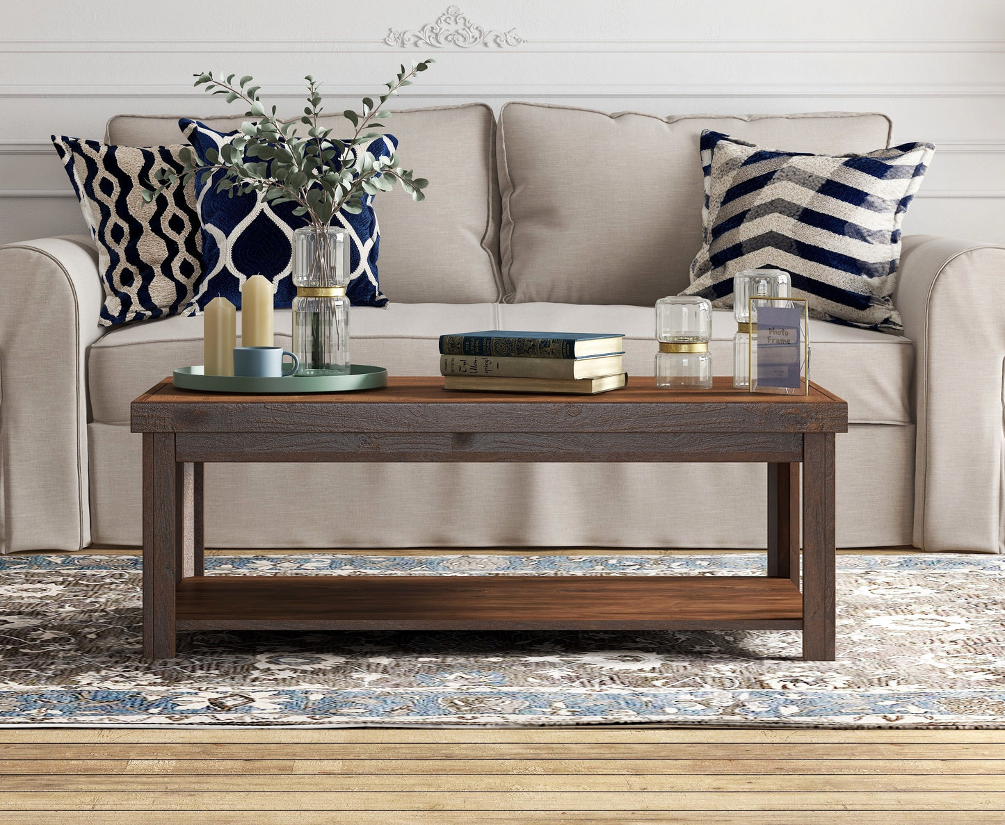 A beige sofa with blue and white pillows sits behind the Couch.com Sausalito Coffee Table, crafted from wood with a dark brown whiskey finish. The table displays books, candles, vases with greenery, and decor atop a patterned rug.