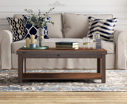 A beige sofa with blue and white pillows sits behind the Couch.com Sausalito Coffee Table, crafted from wood with a dark brown whiskey finish. The table displays books, candles, vases with greenery, and decor atop a patterned rug.