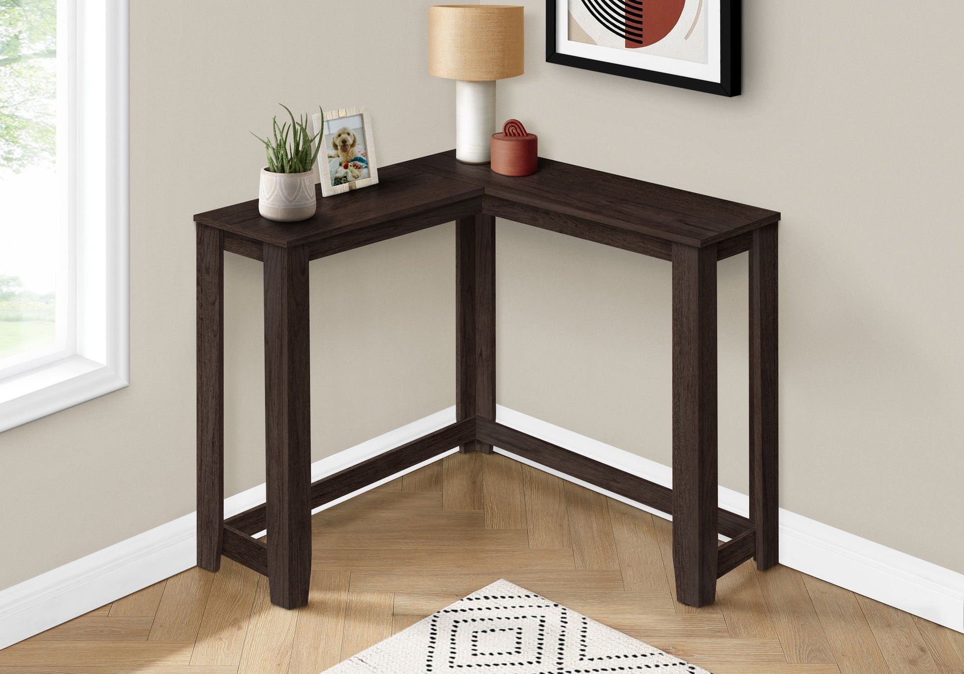 The Corner Accent Console Table For Entryway, Unique Design Contemporary & Modern, is a dark wood L-shaped table set against a beige wall by a window, topped with a lamp, plant, framed photo, and small decorative piece.