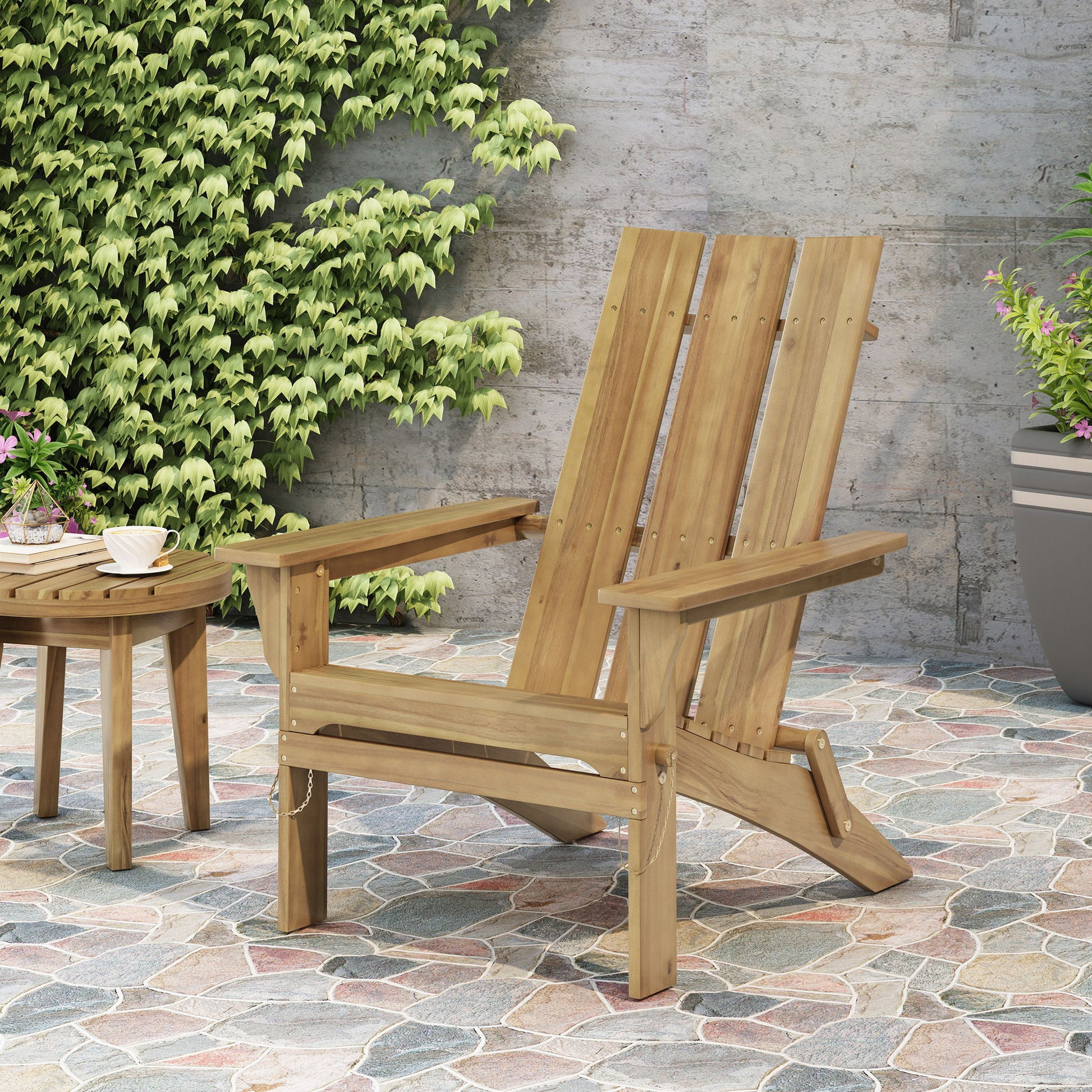 A natural Outdoor Foldable Adirondack Chair with a slat back sits on a stone patio beside a matching round table holding a teacup. Green vines cover the nearby wall, and there's a large potted plant to the right.