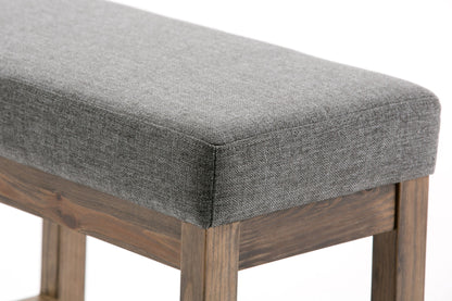 Close-up of the Milltown Upholstered Ottoman Bench, featuring a grey cushion and wooden legs, set against a plain white background.